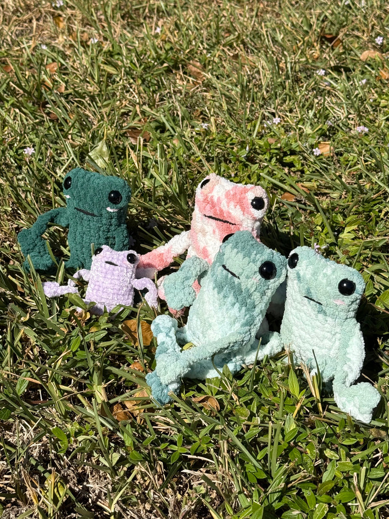 Handmade crochet leggy frog plushies in multiple colors including green, pink, purple and mint with poseable legs displayed on grass