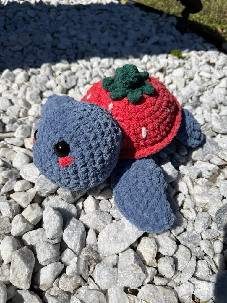 Handmade crochet strawberry turtle plushie with blue body and coral strawberry shell displayed on white decorative rocks in garden setting