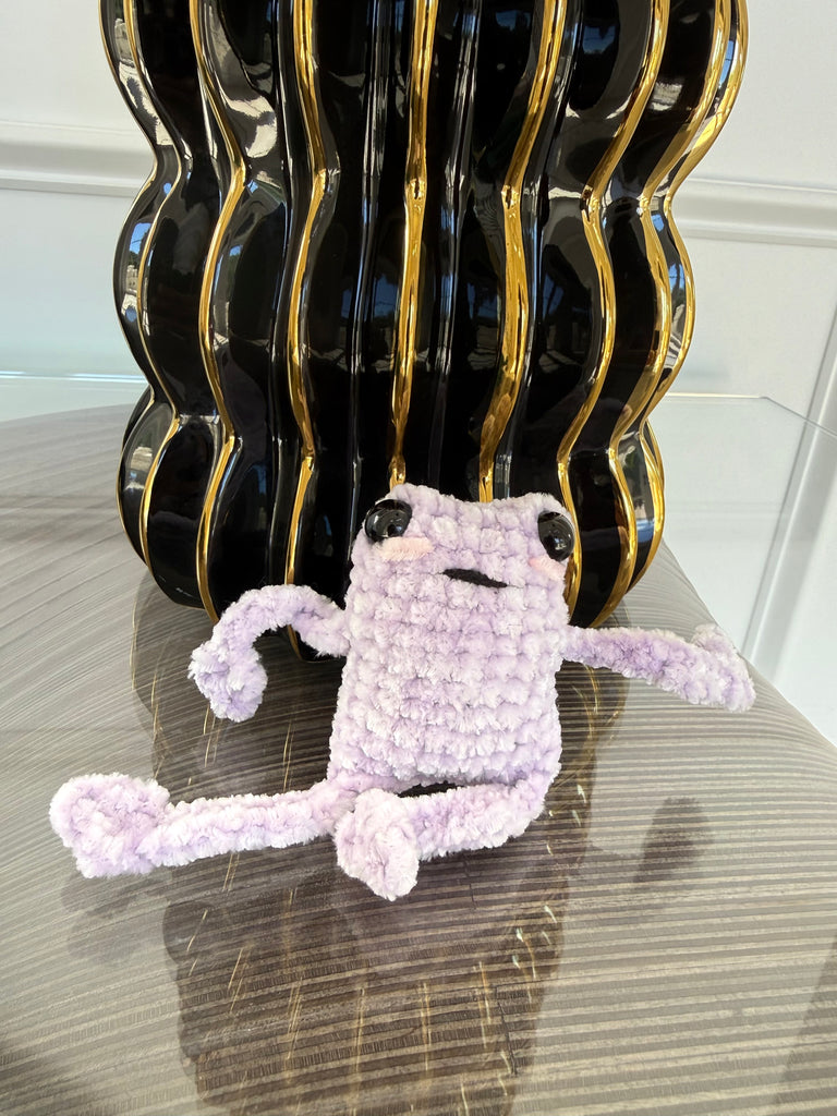 Small purple crochet leggy frog plush toy on a reflective surface with a decorative black and gold vase in the background.