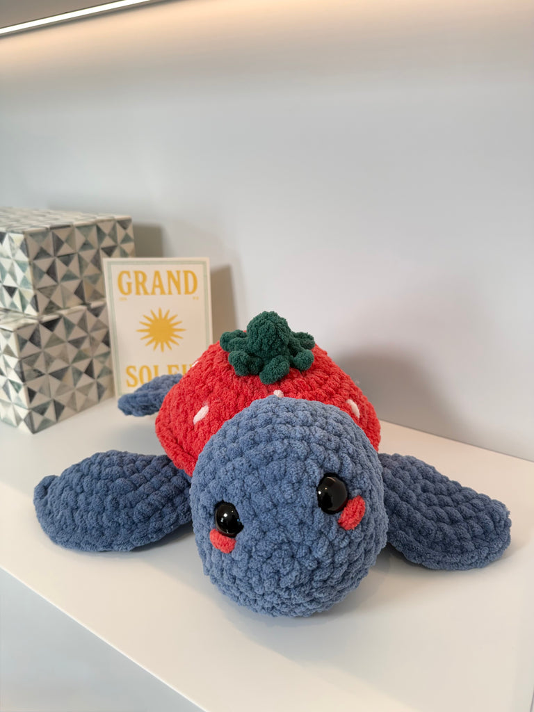 Handmade crochet strawberry turtle plushie in periwinkle blue velvet yarn with red strawberry shell hat and green leaves, featuring embroidered safety eyes and blushing cheeks, displayed on white shelf with home decor