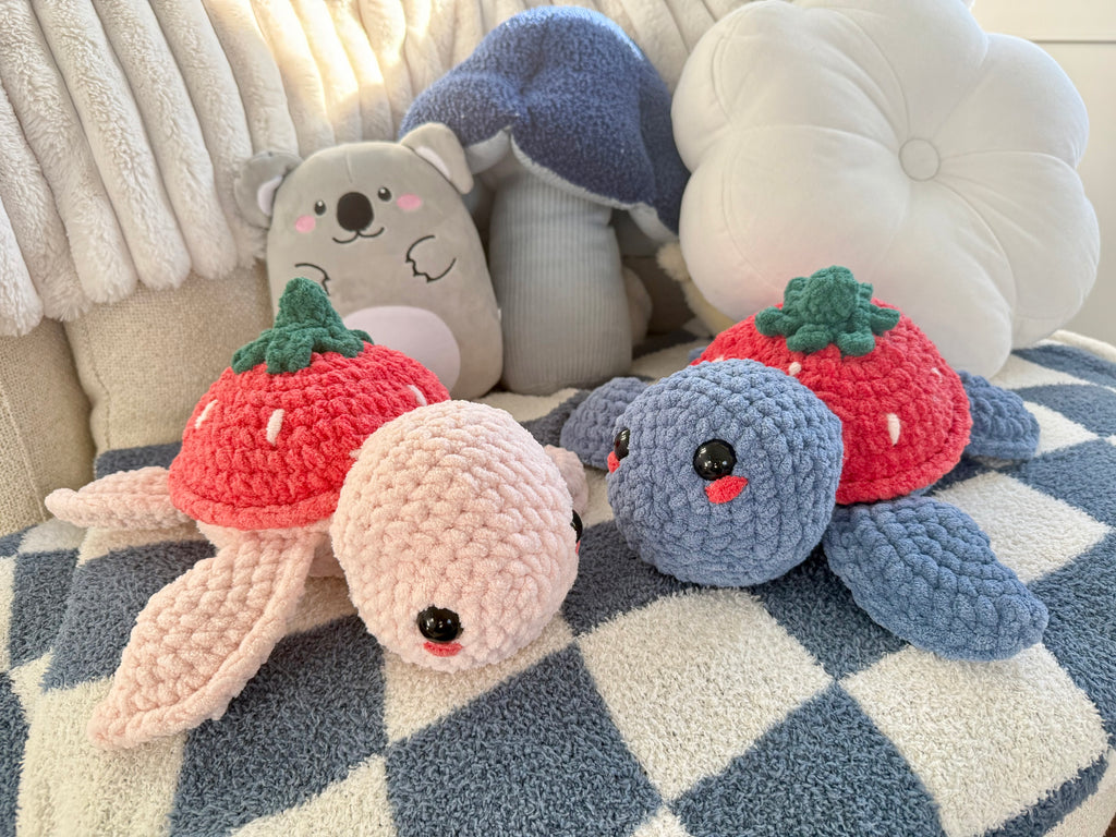 Two handmade crochet strawberry turtle plushies in pink and blue grey sitting together on cozy checkered blanket